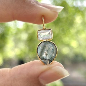 May include: A gold earring featuring a rectangular, clear gemstone above a teardrop-shaped, grey-green stone. The earring is held between fingers, set against a blurred green backdrop.