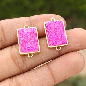 Può includere: Due connettori per gioielli rettangolari caratterizzati da una texture in pietra druzy rosa acceso incastonata in una cornice di metallo color oro lucido. Ogni pezzo include un piccolo anello circolare in alto e in basso per il fissaggio a catene.