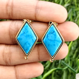 May include: A pair of turquoise gemstone earrings. Each earring features a diamond-shaped, light blue turquoise stone set in a gold-colored metal frame. The earrings are displayed against a green background.