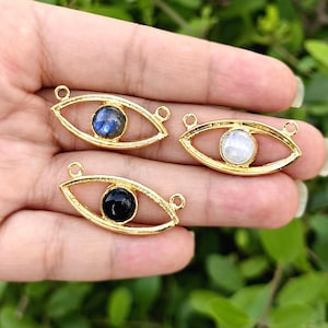 Op de afbeelding: Drie goudkleurige metalen oogvormige sieradenbedels, elk met een kleine ronde edelsteen in het midden. Eén bedel bevat een blauwe iriserende steen, één een witte doorschijnende steen en één een effen zwarte steen.