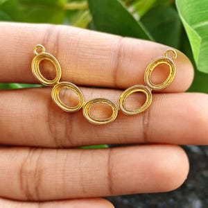 Può includere: Un componente di gioielleria color oro con cinque incastonature ovali collegate in fila. Ogni incastonatura ha un piccolo anello nella parte superiore per il fissaggio. Il componente è presentato su uno sfondo di foglie verdi.