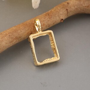 Peut inclure: Un pendentif rectangulaire de couleur dorée avec un bord extérieur texturé à l'aspect organique et un cadre intérieur lisse. La pièce comporte une petite bélière intégrée sur le dessus pour la fixation, avec une finition métallique aux variations subtiles.