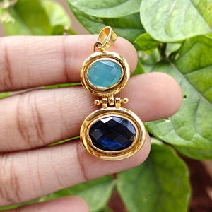 May include: Gold pendant with two oval-shaped gemstones, one teal and one dark blue. The gemstones are set in gold bezels and connected by a gold link. The pendant is held up by a gold loop.
