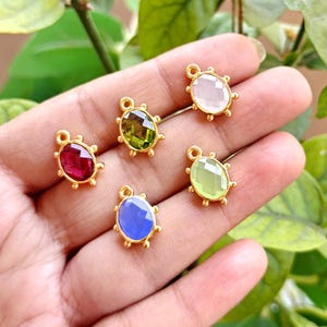 May include: A collection of five gemstone pendants in various colors: red, green, blue, light green, and pink. Each pendant has an oval-shaped gemstone set in a gold-colored metal frame with small, round accents. The pendants are displayed on a hand.
