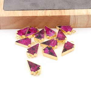 Könnte beinhalten: Eine Sammlung dreieckiger Edelsteinanhänger mit goldfarbenem Rand. Die Edelsteine sind ein leuchtendes Pink mit dunklen lila und schwarzen Einschlüssen. Jeder Anhänger hat eine kleine Schlaufe zum Befestigen.