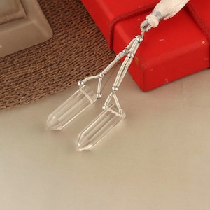 May include: Clear quartz crystal pendants with silver-colored wire wrapping and a white tassel. The pendants are rectangular with pointed ends. The background includes a red box and a woven brown surface.