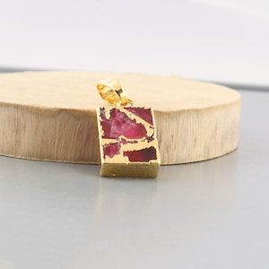 Puede incluir: Un colgante dorado con una piedra rectangular de color rubí. La piedra está engastada en un marco dorado y tiene un lazo dorado para una cadena. El colgante se muestra sobre una superficie de madera.