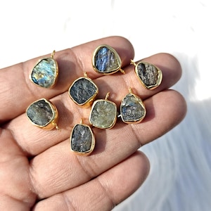 Pode incluir: Uma coleção de pingentes banhados a ouro, cada um com uma pedra preciosa bruta e iridescente. As pedras exibem uma variedade de cores, incluindo azuis, verdes e cinzas. Os pingentes são segurados em uma mão, mostrando suas formas e texturas únicas.