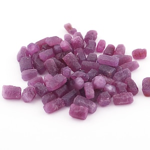 Pode incluir: Uma pilha de pedras preciosas de rubi brutas e naturais em vários tons de rosa e roxo. As pedras preciosas têm formato irregular e aparência ligeiramente translúcida, com algumas pedras mais escuras e opacas misturadas. O fundo é branco.