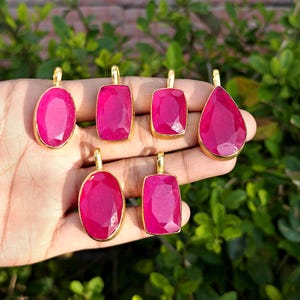 May include: A collection of six ruby pendants in various shapes: oval, rectangular, and teardrop. Each pendant features a vibrant pink gemstone set in a gold-colored bezel. The pendants are displayed against a natural green background.