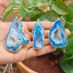 May include: Three agate geode pendants with blue and white crystalline centers and gold-toned metallic edges. Each pendant features a small metal loop at the top for attachment, showcasing natural stone variations in shape, size, and blue color intensity.