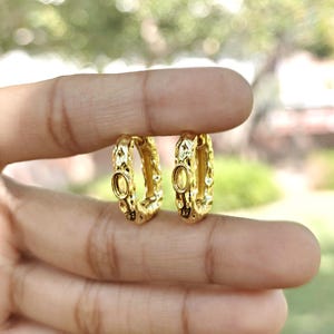 May include: A pair of gold-colored hoop earrings. Each earring has an oval-shaped setting in the center and a textured, patterned design around the hoop. The earrings are held between fingers against a blurred background.