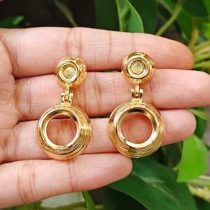 May include: A pair of gold-toned drop earrings featuring a small circular stud top connected to a larger, hollow circular pendant. The metal has a polished, reflective finish with subtle concentric ring detailing around the outer edges of the circular components.