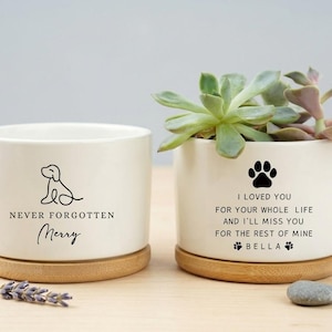 May include: Two white ceramic planters with wooden bases. One has a dog outline and the text "NEVER FORGOTTEN Merry." The other has a paw print and the text "I LOVED YOU...BELLA." One planter holds succulents.