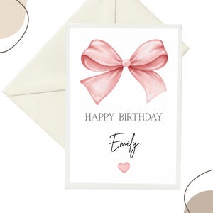 Può includere: Biglietto di compleanno con un'illustrazione ad acquerello di un fiocco rosa. La carta dice "HAPPY BIRTHDAY Emily" con un piccolo cuore rosa sotto il nome. Una busta bianco sporco è dietro la carta.