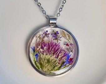 Op de afbeelding: Een zilverkleurige hangerketting met een heldere, ronde harscharm. De charm bevat gedroogde bloemen in paars, blauw en groen. De ketting is ook zilverkleurig. De hanger heeft een diameter van ongeveer 3 cm.