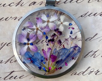 Op de afbeelding: Een ronde, zilverkleurige hanger met een helder harsmiddel gevuld met gedroogde paarse, witte en blauwe bloemen. De ketting heeft een ketting met twee kleine, roze edelsteenaccenten.