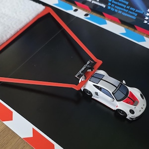 Op de afbeelding: Een witte en rode speelgoed raceauto met een zwarte spoiler staat op een zwart oppervlak. Een rood driehoekig frame en een witte doek zijn ook aanwezig. Het oppervlak heeft rood-witte markeringen.