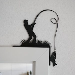 Peut inclure: Silhouette noire en métal d'un pêcheur attrapant un poisson. La pièce décorative représente un pêcheur avec une canne à pêche, de l'herbe et un poisson à l'hameçon. Le design est monté sur un cadre de porte blanc.