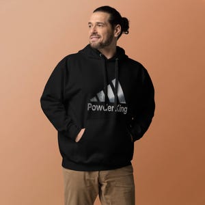 Può includere: Felpa con cappuccio nera con tasca frontale e coulisse. La felpa presenta una grafica di montagna bianca e grigia con il testo "Powder King" in un font stilizzato. Il modello indossa pantaloni color cachi.