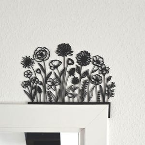 Könnte beinhalten: Schwarze Metall-Blumenwandkunst mit verschiedenen Blumensilhouetten. Das dekorative Stück ist über einem weißen Türrahmen angebracht und bildet einen Kontrast zur weißen Wand. Das Design umfasst detaillierte Umrisse verschiedener Blumensorten.