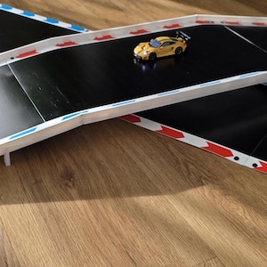 Könnte beinhalten: Ein gelbes Spielzeug-Rennauto auf einer schwarzen Rennstrecke mit weißen, roten und blauen Akzenten. Die Strecke ist erhöht und kreuzt einen anderen Abschnitt. Die Strecke befindet sich auf einer Holzoberfläche.