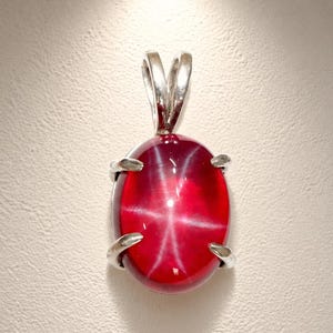 May include: Oval-shaped ruby pendant with a star effect. The deep red gemstone is set in a silver metal frame with a loop for a chain. The pendant is displayed against a white background.