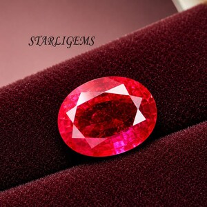 May include: An oval-cut, vibrant red gemstone with a faceted surface, displaying internal light reflections. The stone is positioned on a dark maroon velvet-textured surface. The text STARLIGEMS is printed in black in the upper left corner.