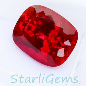Op de afbeelding: Een gefacetteerde, kussenvormige, dieprode edelsteen rust in een witte doos. De edelsteen heeft een briljant, reflecterend oppervlak. De tekst "StarliGems" is zichtbaar in de rechter benedenhoek.