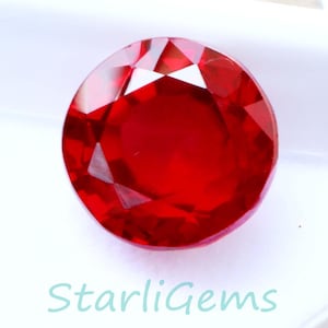 May include: A round, faceted, bright red gemstone, presented in a white plastic case. The gem has a brilliant cut, reflecting light. The text "StarliGems" is printed in a light blue colour on the case.