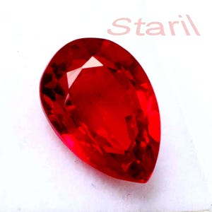 May include: A faceted, pear-shaped red gemstone. The gem is displayed on a white surface, highlighting its brilliant cut and intense colour. The word "Staril" is faintly visible in the background.