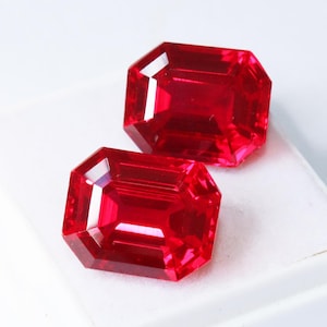 Puede incluir: Dos gemas rojas profundas, talladas en forma rectangular y facetadas. Las gemas se exhiben sobre una superficie blanca, mostrando sus superficies pulidas y brillantes y sus reflejos internos. Las gemas son de un color rojo vibrante y saturado.