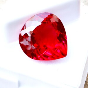 May include: A faceted, heart-shaped, vibrant red gemstone. The gem is set in a white display case, showcasing its brilliant cut and intense color. The gemstone's facets reflect light, creating a sparkling effect. This item is suitable for jewelry making.