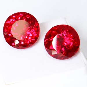 Könnte beinhalten: Zwei runde, facettierte, leuchtend rote Edelsteine. Die Edelsteine werden auf einer weißen Oberfläche präsentiert, die ihren Brillantschliff und ihre lebendige Farbe zur Geltung bringt. Bei den Edelsteinen handelt es sich wahrscheinlich um Rubine.