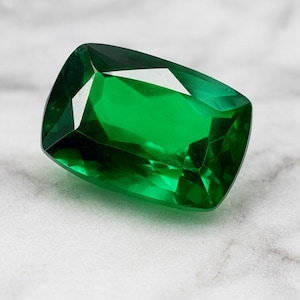 May include: A radiant, emerald-cut gemstone in a rich green shade. The gem's facets catch the light, producing a brilliant sparkle. It rests on a white marble surface, accentuating its colour and clarity. The gem is approximately 2 cm in length.