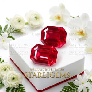 Può includere: Due gemme rosso vibrante, taglio smeraldo, appoggiate su una scatola rettangolare bianca. La scatola riporta il testo STARLIGEMS PREMIUM GEMS & JEWELRY. La composizione è circondata da fiori bianchi e foglie di felce verdi su sfondo bianco.