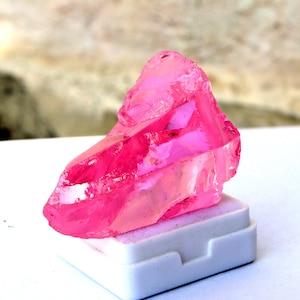 May include: A vibrant, raw, pink crystal gemstone, resting on a white base. The crystal has a rough, irregular shape with a translucent quality, showcasing its natural beauty. The gemstone is a striking shade of hot pink.