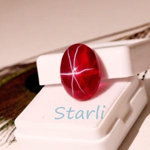 May include: A polished, oval-shaped, red gemstone with a star-like pattern of white lines. The gemstone is displayed on a white surface, with the word "Starli" in light blue script. The gem is a cabochon cut.