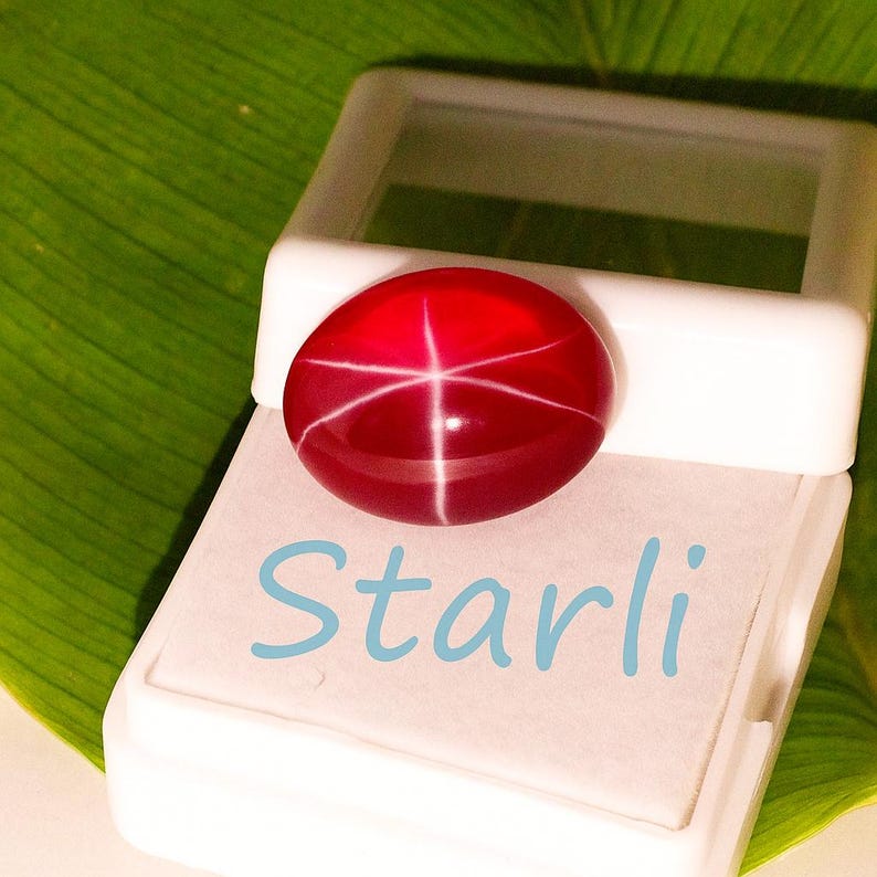 May include: A polished, oval-shaped, red gemstone with a star-like pattern, displayed in a white box. The word "Starli" is written in light blue on the box. The background features a green leaf.