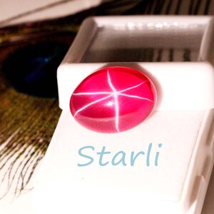 May include: A vibrant, oval-shaped, ruby-red gemstone with a star-like pattern of white lines. The gem is displayed on a white surface, with the word "Starli" in light blue script below it. The image suggests a jewelry or gemstone product.