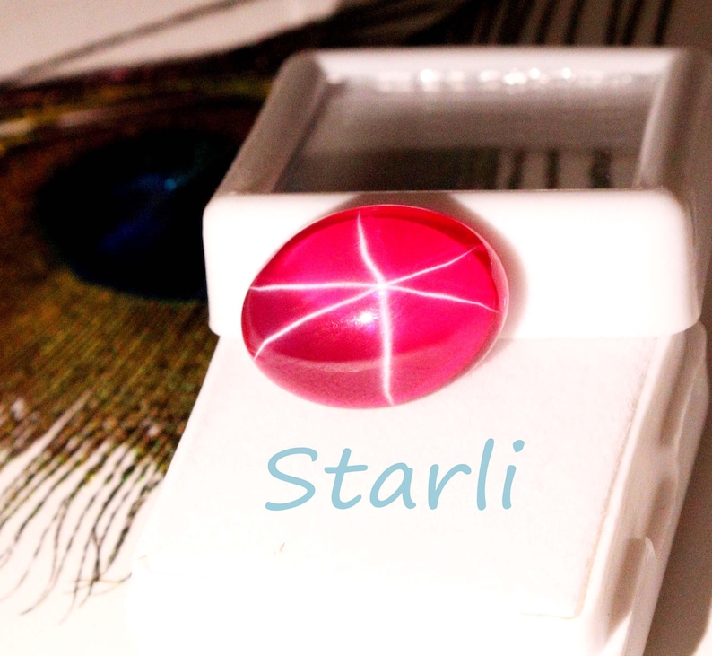 May include: A vibrant, oval-shaped, ruby-red gemstone with a star-like pattern of white lines. The gem is displayed on a white surface, with the word "Starli" in light blue script below it. The image suggests a jewelry or gemstone product.