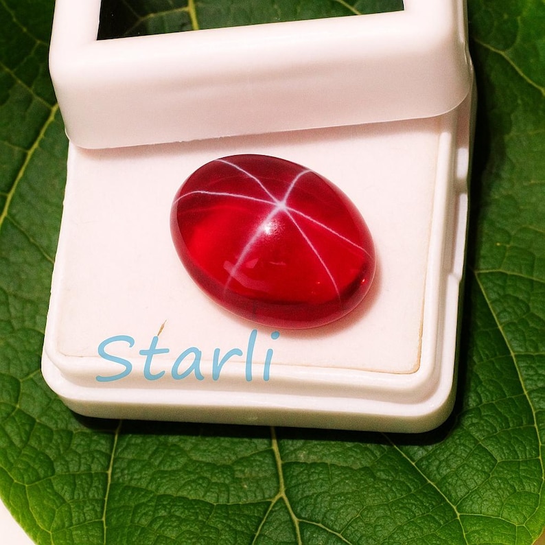 May include: A polished, oval-shaped, red gemstone with a six-rayed star pattern, displayed in a white box. The gemstone is set against a green leaf background. The word "Starli" is written in light blue at the bottom.