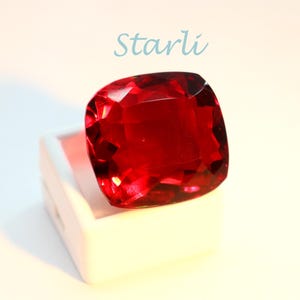 May include: A large, faceted, cushion-cut red gemstone, presented in a white box. The gem has a deep, rich colour and a brilliant shine. The word "Starli" is written in light blue script above the gem.