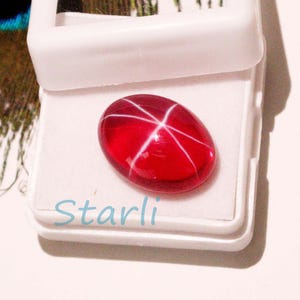 May include: A vibrant red oval gemstone with a six-rayed star pattern, displayed in a white box. The gemstone has a polished, glossy surface. The word "Starli" is printed in light blue on the box. This image showcases a precious stone.