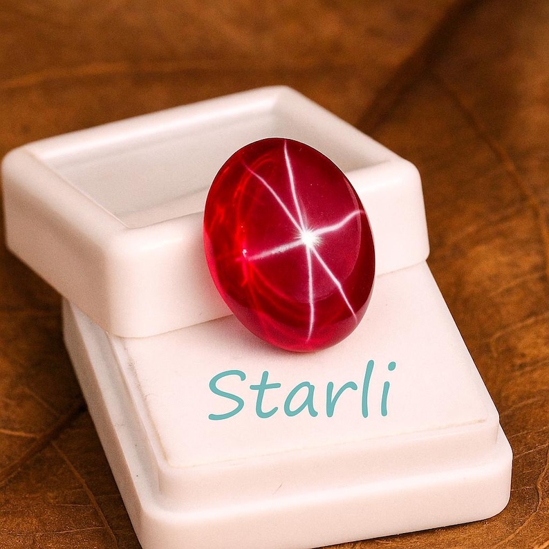 May include: A polished, oval-shaped, red gemstone with a star-like pattern of white lines. The gemstone rests on a white display case, with the word "Starli" in teal script. The background is a warm, brown tone.