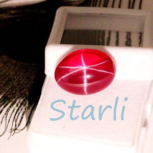 May include: A vibrant red gemstone with a star-like pattern, set against a white background. The word "Starli" is written in light blue script below the gem. The gemstone is oval-shaped and appears to be in a display case.