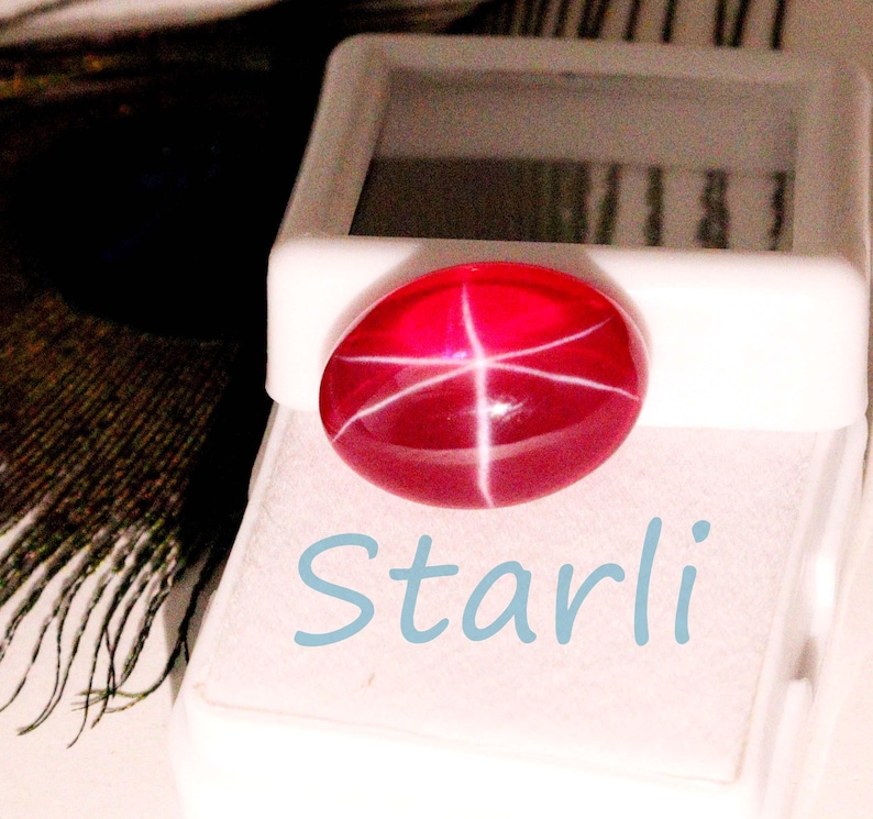 May include: A vibrant red gemstone with a star-like pattern, set against a white background. The word "Starli" is written in light blue script below the gem. The gemstone is oval-shaped and appears to be in a display case.