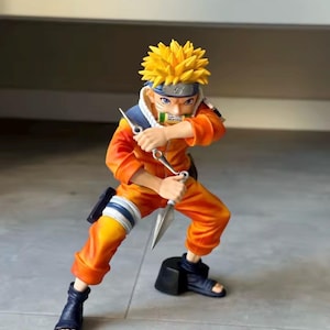 May include: A figurine of a character in an orange jumpsuit with yellow hair and blue accents. The figure is holding two silver kunai knives and is in a dynamic pose, ready for action. The figure is wearing black sandals.