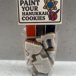 Peut inclure: Un sac en plastique transparent contenant des biscuits de Hanoucca avec du glaçage blanc, dont un menorah, un dreidel et d'autres formes. L'étiquette indique "KOSHER PAINT YOUR HANUKKAH COOKIES" avec une étoile de David et une palette de peinture.