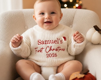 May include: A cream-colored long-sleeve knit sweater featuring red embroidered text that reads Samuel's First Christmas 2026, accompanied by a small red and white Santa hat icon positioned above the text on the front of the garment.